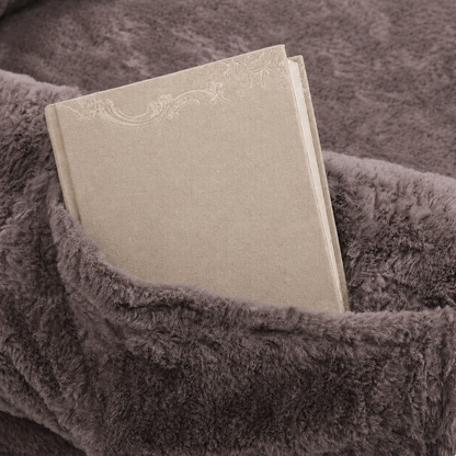 A beige fabric-covered book or notebook with a subtle embossed floral design, tucked into a plush, textured gray blanket.
